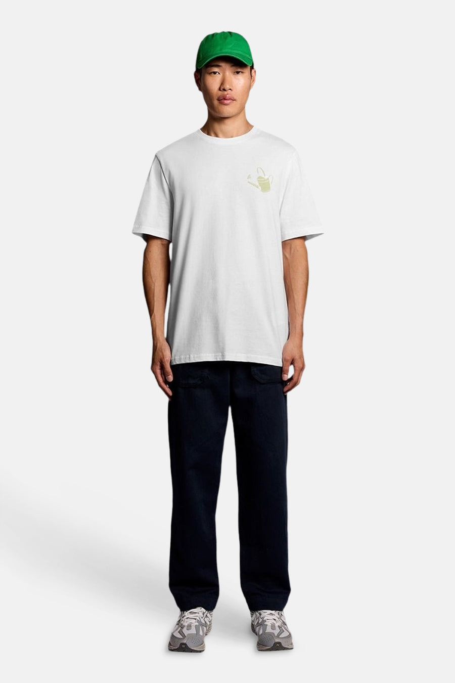 T-shirt blanc de LYLE & SCOTT avec motif d'arrosoir vert clair, porté par un homme avec une casquette verte, un pantalon bleu marine et des baskets grises.