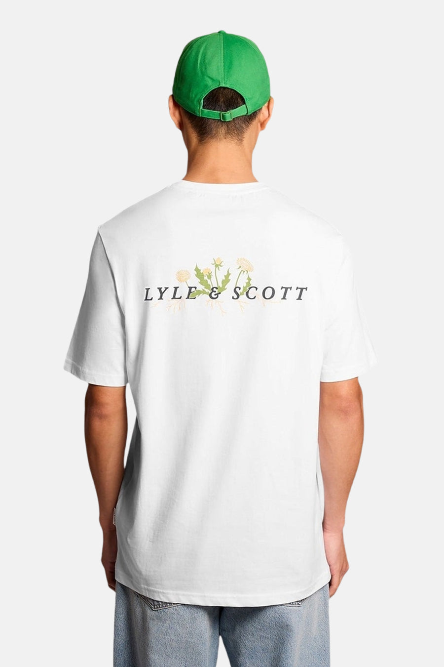 Wit T-shirt van LYLE & SCOTT met bloemen "LS"-logo, gedragen met een lichte spijkerbroek, loafers en een groene pet.