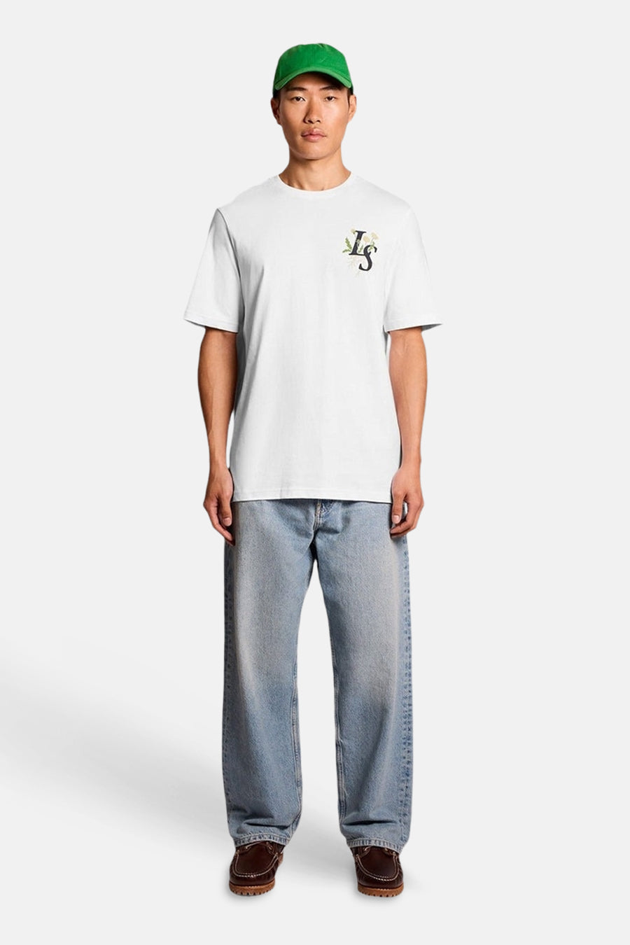 T-shirt blanc de LYLE & SCOTT avec logo "LS" floral, porté avec un jean clair, des mocassins et une casquette verte.