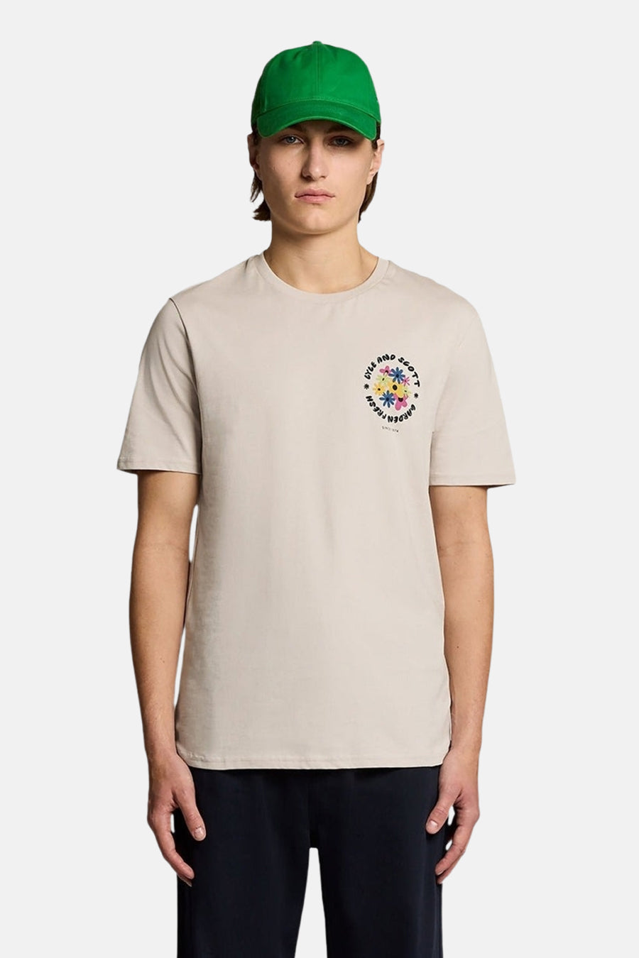 Ecru T-shirt van LYLE & SCOTT met meerkleurig bloemenpatroon op de borst, gedragen door een man in een donkere broek en een groene pet.