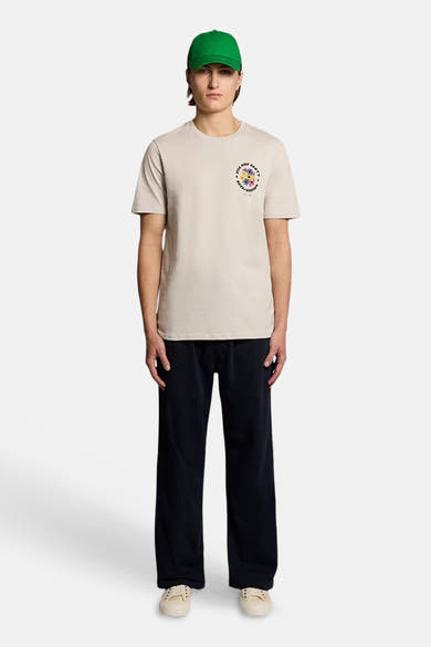 T-shirt écru de LYLE & SCOTT avec petit imprimé floral coloré sur la poitrine, porté avec pantalon large et casquette verte.