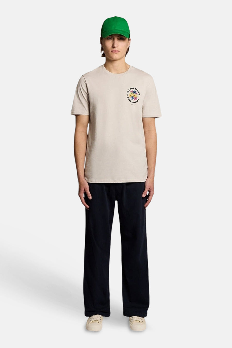 T-shirt écru de LYLE & SCOTT avec petit imprimé floral coloré sur la poitrine, porté avec pantalon large et casquette verte.