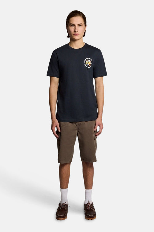 T-shirt bleu de LYLE & SCOTT avec motif circulaire coloré sur la poitrine, porté avec un short cargo marron et des chaussures bateau.