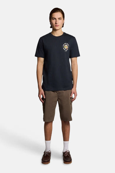 T-shirt bleu de LYLE & SCOTT avec motif circulaire coloré sur la poitrine, porté avec un short cargo marron et des chaussures bateau.