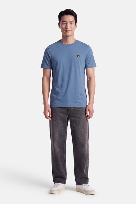 T-shirt bleu à manches courtes par LYLE & SCOTT, col rond, petit logo brodé sur poitrine, porté avec jeans gris et baskets blanches, vue de face.