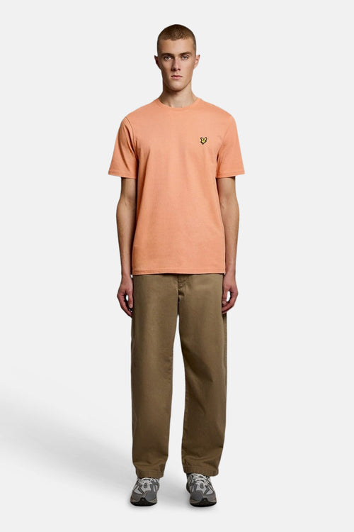 T-shirt à manches courtes orange de LYLE & SCOTT avec emblème aigle jaune, porté avec un pantalon marron et des baskets grises.