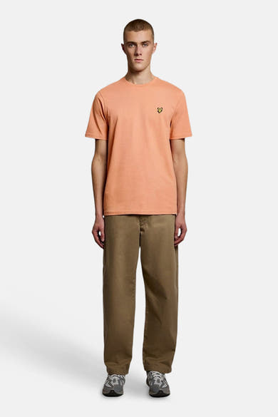 Oranje T-shirt met korte mouwen van LYLE & SCOTT met geel adelaarslogo, gedragen met een bruine broek en grijze sneakers.