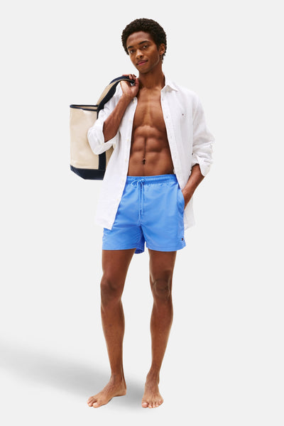 Short de bain bleu de Tommy Hilfiger porté avec une chemise blanche ouverte et un sac de plage.