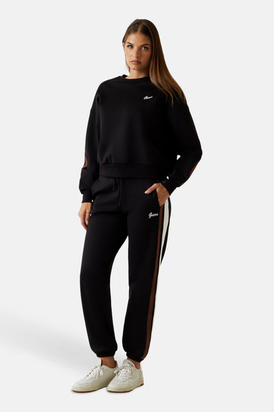 Jogging noir de Guess avec une bande marron et blanche sur le côté, porté avec des baskets blanches.