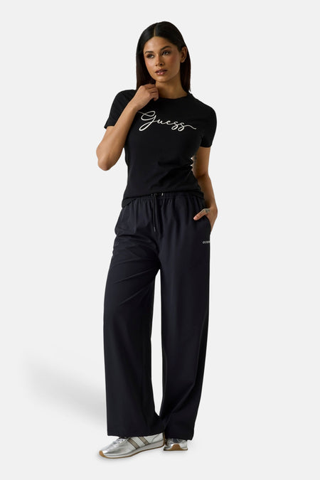 T-shirt noir à manches courtes de Guess avec logo script blanc, porté avec un pantalon noir et des baskets argentées.