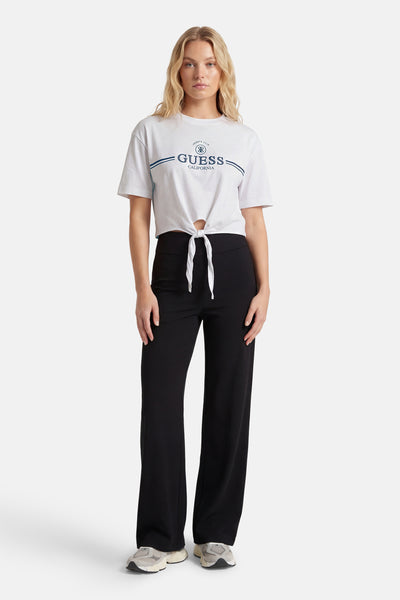 T-shirt blanc à manches courtes de Guess, noué à la taille, porté avec un pantalon noir ample et des baskets grises.
