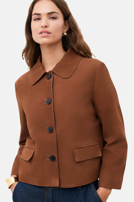 Veste courte brune de CAROLL avec col rond, boutons foncés et poches à rabat, portée avec une boucle d'oreille et un bracelet manchette dorés.