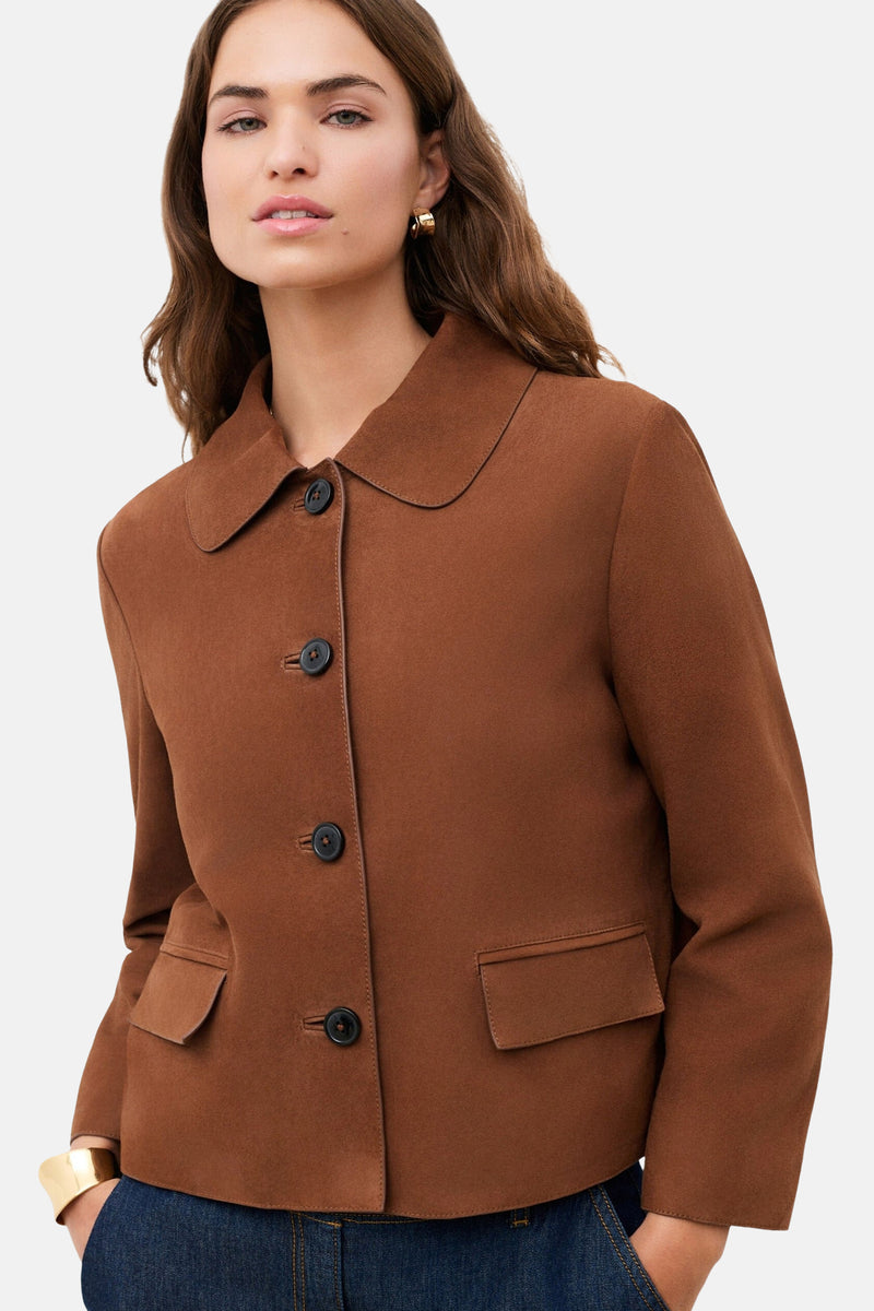 Veste courte brune de CAROLL avec col rond, boutons foncés et poches à rabat, portée avec une boucle d'oreille et un bracelet manchette dorés.