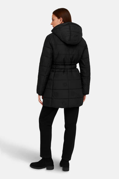 Zwarte lange jas van Guess, met capuchon en riem, gedragen met een zwarte broek en enkellaarsjes.
