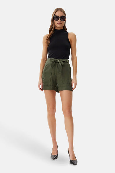 Korte groene Guess-short met hoge taille en strikceintuur, gedragen met een zwart topje en hakken.