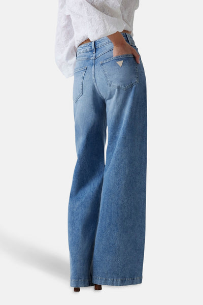 Wijde spijkerbroek in middinblauw denim van Guess, met een hand in de achterzak, gedragen met een witte blouse.