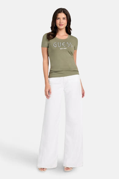 T-shirt kaki à manches courtes de Guess, porté avec un pantalon blanc ample et des sandales.