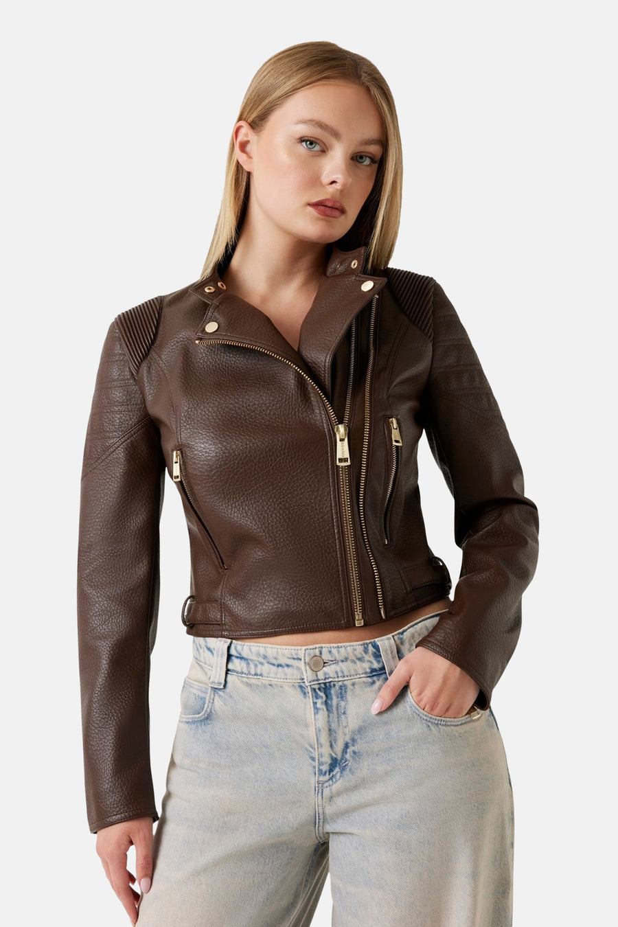 Veste en simili cuir brun de Guess, avec fermeture éclair dorée et détails côtelés aux épaules, portée avec un jean clair.
