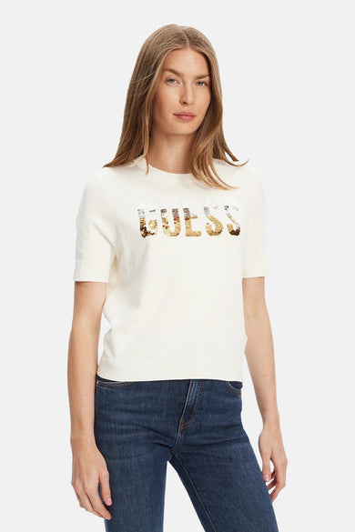 Pull à col rond écru de Guess avec motif logo pailleté sur le devant, porté avec un jean bleu foncé.

