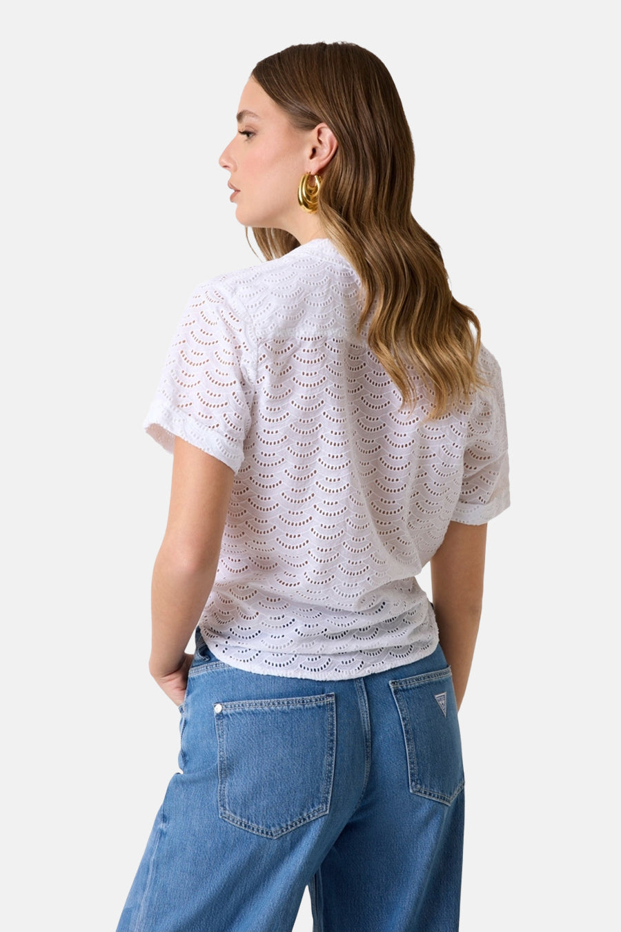 Wit blouse met korte mouwen van Guess® met golvend ajourpatroon, van achteren gedragen met een blauwe spijkerbroek en gouden oorbellen.