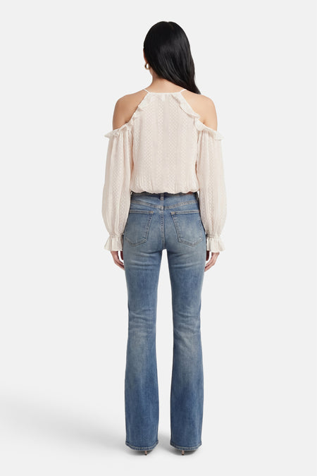 Blouse à manches longues écru de Guess, épaules dénudées et détails volantés, portée avec un jean bleu taille haute.
