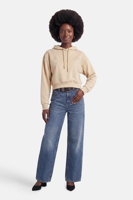 Sweat à capuche beige court de Guess® avec motif de feuilles en relief, porté sur un jean bleu et une ceinture noire.