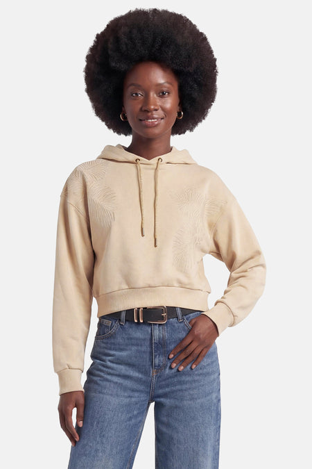 Sweat à capuche beige de Guess, avec de subtils motifs de feuilles de palmier brodés, coupe courte, porté avec un jean.