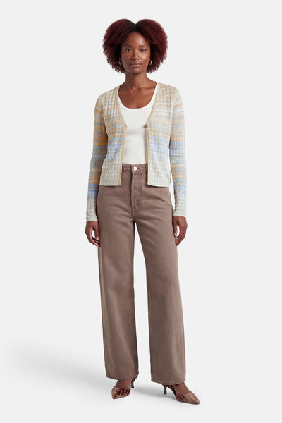 Gilet multicolore par Guess, à rayures horizontales aux tons beige, orange et bleu, porté sur un débardeur blanc et un pantalon marron.