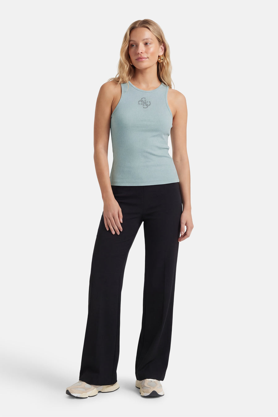 Top sans manches vert de Guess, avec un logo décoratif et une texture côtelée, porté avec un pantalon noir et des chaussures de sport.
