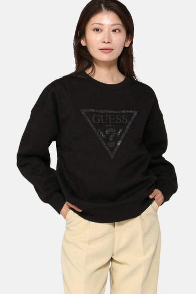 Zwarte Guess sweater met ronde hals, met strass driehoekig logo op de voorkant, gedragen met een beige broek.