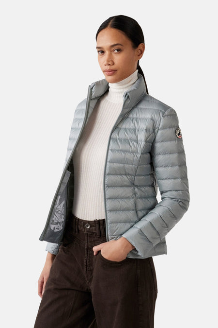 Veste de pluie grise par JOTT, matelassée et portée sur un pull blanc à col roulé et un pantalon marron.