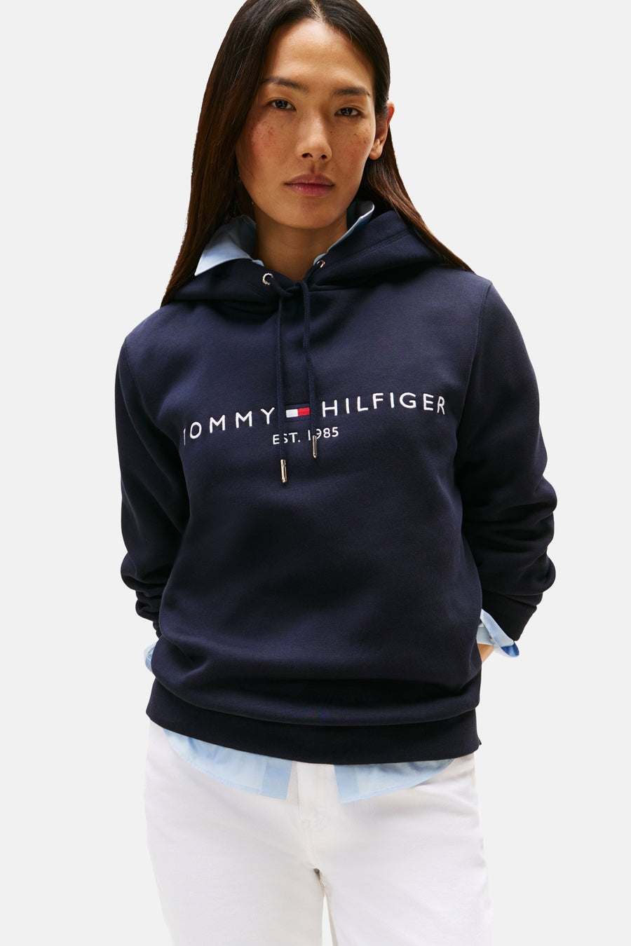 Donkerblauwe hoodie van TOMMY HILFIGER, met wit logo op de voorkant, gedragen over een lichtblauw overhemd en een witte broek.