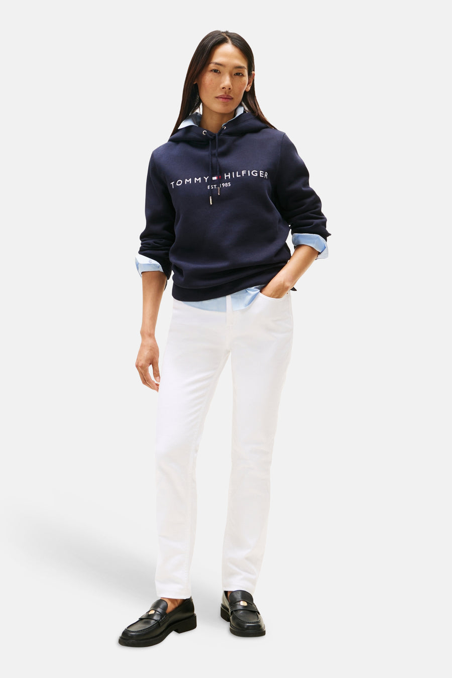 Sweat à capuche bleu marine de TOMMY HILFIGER, avec logo blanc sur le devant, porté sur une chemise bleu clair et un pantalon blanc.