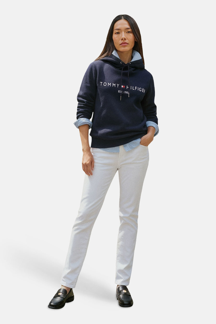 Blauwe hoodie van TOMMY HILFIGER, gedragen over een licht overhemd, met een witte broek en loafers.