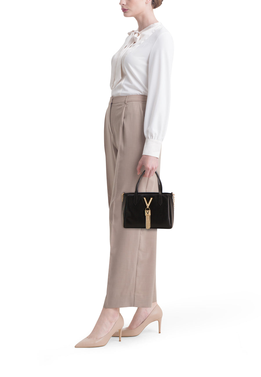 Sac à main noir de VALENTINO BAGS avec logo doré, porté avec chemisier blanc et pantalon beige.