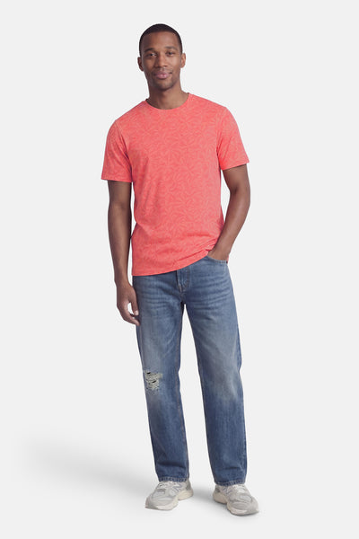 Coral short-sleeve t-shirt with subtle floral pattern, worn by model in full-length lifestyle view with blue jeans and sneakers.