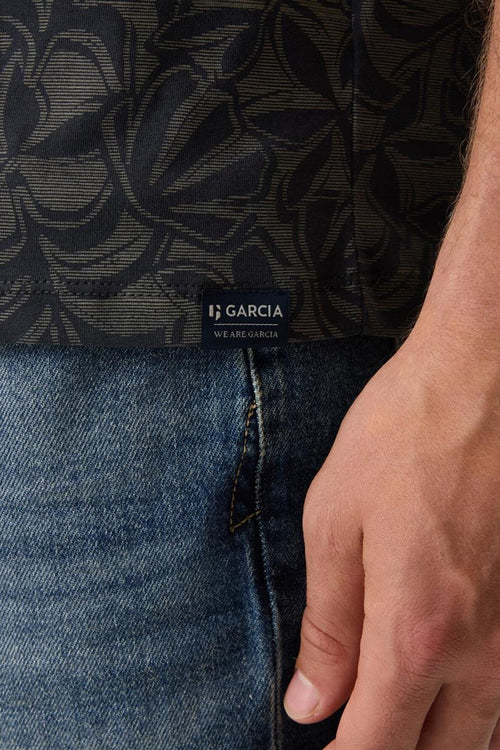 T-shirt bleu à motif de feuilles, de Garcia, porté avec un jean bleu et une main visible.