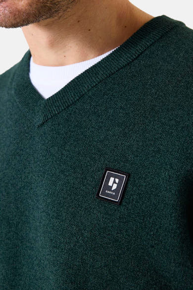 Groene V-hals trui van GARCIA, versierd met een logo-embleem op de borst, gedragen over een wit T-shirt.