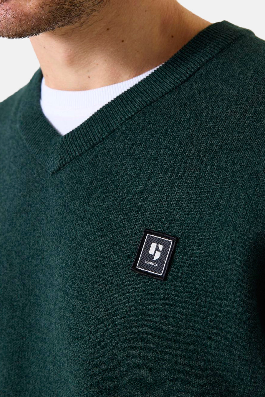 Groene V-hals trui van GARCIA, versierd met een logo-embleem op de borst, gedragen over een wit T-shirt.
